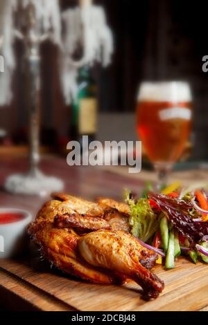 Pollo fritto succoso con verdure ed erbe e un bicchiere di birra leggera sul tavolo nel bar in colori scuri Foto Stock