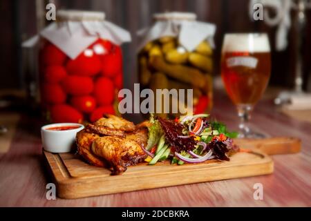 Pollo fritto succoso con verdure ed erbe e un bicchiere di birra leggera sul tavolo nel bar in colori scuri Foto Stock