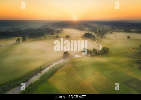 Paesaggio all'alba - un piccolo fiume nella nebbia con alberi e cespugli tra i verdi prati estivi - alba gialla sul prato Foto Stock