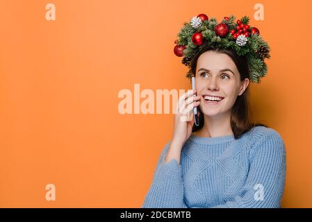 Bella donna sorridente indossa la corona di Natale, parlando sul telefono cellulare che conduce una piacevole conversazione, isolato su studio arancione Foto Stock