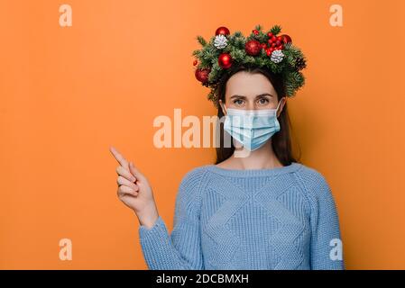 Scatto orizzontale di giovane donna che indossa i punti della maschera medica con la mano da parte, mostra lo spazio di copia per la pubblicità, indossa la corona di vacanza di abete rosso Foto Stock