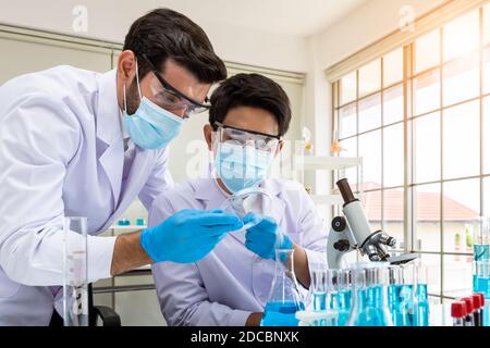 Due ricercatori asiatici e arabi maschi che lavorano in laboratorio, conducendo studio sostanza a rischio biologico con apparecchiature scientifiche e microscopio Foto Stock