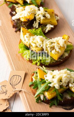 Panini tostati con formaggio brie, zucca al forno e insalata, rucola e mozzarella, deliziosi spuntini vegetali su tavola di legno Foto Stock