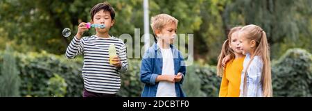 Ragazzo asiatico soffiare bolle di sapone vicino sorridenti amici all'aperto, striscione Foto Stock