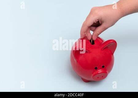 Primo piano di una mano di un bambino mette una moneta in un banco di piggy rosso su uno sfondo blu, vista dall'alto, spazio di copia. Concetto di risparmio di denaro Foto Stock