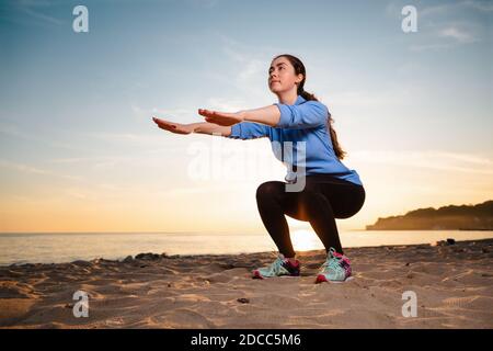 Una giovane donna in abbigliamento sportivo si allena facendo squadre. Sullo sfondo, il tramonto, la spiaggia sabbiosa e l'oceano. Vista dal basso. Foto Stock