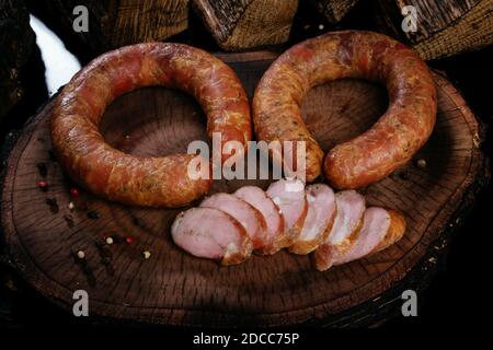 Due anelli di salsiccia affumicata su una tavola, sullo sfondo di legna da ardere bruciata. Diversi pezzi di salsiccia a fette. Foto Stock