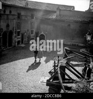 Una romantica passeggiata di coppia che tiene le mani attraverso una soleggiata piazza acciottolata a Roma, passando davanti ad alcuni giganteschi croci, mentre un fascio di sole si riversa giù. Foto Stock