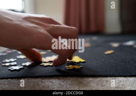 Primo piano di teenager che fa un puzzle a casa su un tappeto di puzzle di stoffa nera sul tappeto del pavimento circa per raccogliere un singolo pezzo di puzzle Foto Stock