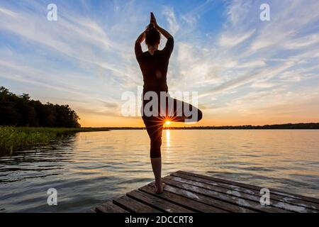 Donna che pratica la postura in piedi Vrikshasana / posa albero, una zampe di equilibrio asana della hatha medievale yoga sul molo al lago al tramonto Foto Stock