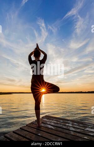 Donna che pratica la postura in piedi Vrikshasana / posa albero, una zampe di equilibrio asana della hatha medievale yoga sul molo al lago al tramonto Foto Stock