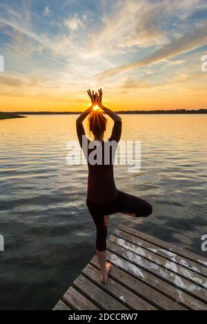 Donna che pratica la postura in piedi Vrikshasana / posa albero, una zampe di equilibrio asana della hatha medievale yoga sul molo al lago al tramonto Foto Stock