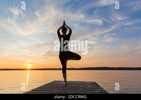 Donna che pratica la postura in piedi Vrikshasana / posa albero, una zampe di equilibrio asana della hatha medievale yoga sul molo al lago al tramonto Foto Stock