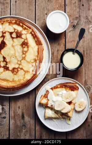 Frittelle sottili, crepes con banane e latte condensato in un piatto su un tavolo di legno Foto Stock