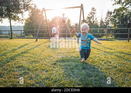 Un ragazzo di un anno che corre a piedi nudi nel parco giochi su Un bel giorno di primavera - carino ragazzino che lo prende Primi passi al tramonto - imparare a camminare conce Foto Stock