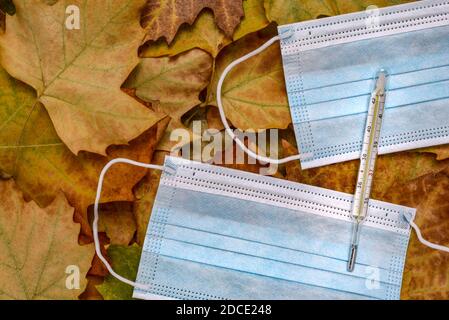 Maschere facciali blu e termometro in vetro per misurare la temperatura del corpo umano su foglie secche dell'autunno. Foto Stock
