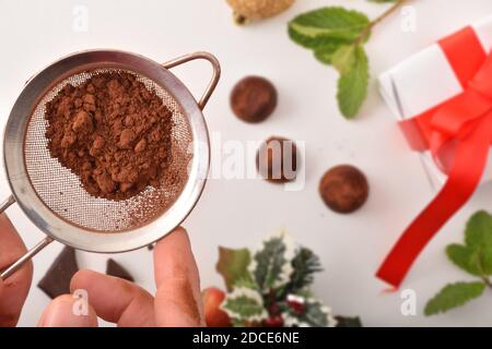 Mani versando polvere di cacao con un setaccio sopra tartufi di cioccolato su panca da cucina decorata con ornamenti di regalo e di natale. Vista dall'alto. Orizzontale Foto Stock