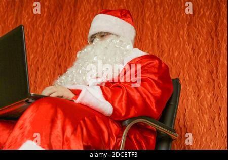 Babbo Natale è seduto nella sua stanza su una sedia con un computer portatile in mano. Foto Stock