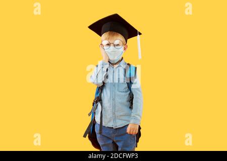 Bambino ragazzo studente di scuola elementare in cappello laureato con un zaino sulla schiena con una maschera protettiva che torna indietro a scuola dopo covid-19 quaranti Foto Stock