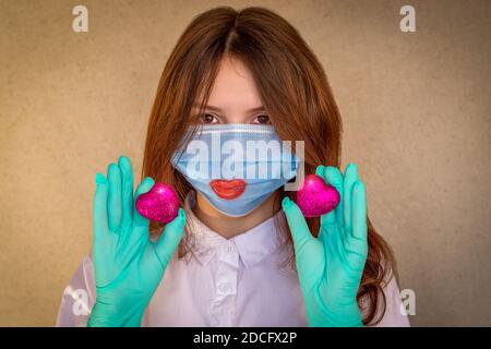 Una ragazza adolescente che indossa una maschera medica e guanti, tiene due cuori scarlatto davanti a lei. Le labbra rosse sono dipinte sulla maschera. Messa a fuoco selettiva Foto Stock