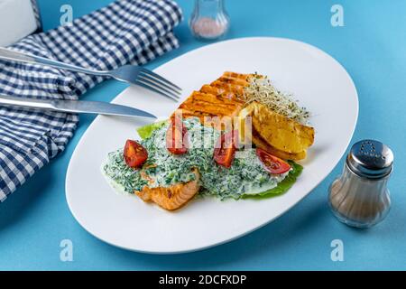Bistecca di salmone alla griglia. Un pezzo di pesce rosso su un piatto bianco e uno sfondo blu sul tavolo. Pesce con salsa, pomodori, limone. Menu del ristorante Foto Stock