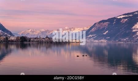 Zell am See in inverno sera. Vista sul lago Zell, città, montagne e neve con riflessi in acqua. Città alpina al crepuscolo viola. Famosa stazione sciistica Foto Stock