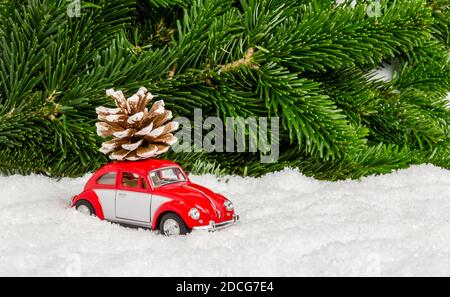 Fulda, Germania - 24 ottobre 2020: Piccola auto rossa giocattolo nella neve con cono di pino davanti all'abete. Consegna in vacanza invernale o concetto di viaggio Foto Stock