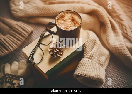 Una tazza di caffè caldo con aroma panna si trova sopra una pila di vecchi libri, accanto ad esso sono bicchieri neri e un cono di abete, e accanto ad esso è una sudorazione calda Foto Stock