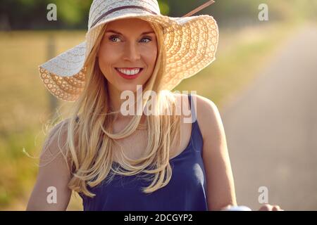 Bella donna bionda di mezza età in un ampio cappello di paglia brimmed e la parte superiore blu dell'estate sorridente alla macchina fotografica come lei sguardi da parte su una strada rurale soleggiata Foto Stock