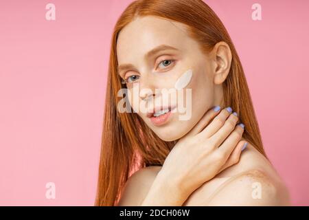 Closeup bellezza ritratto di redhead caucasian bellissima femmina l'applicazione di crema idratante viso o primer e guardando la telecamera su sfondo rosa. Y Foto Stock
