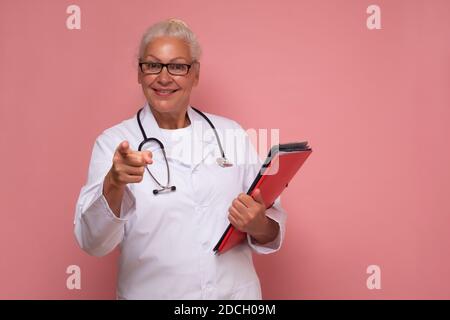Donna dottore senior sorridente e indicando con l'indice dito isolato su sfondo rosa Foto Stock