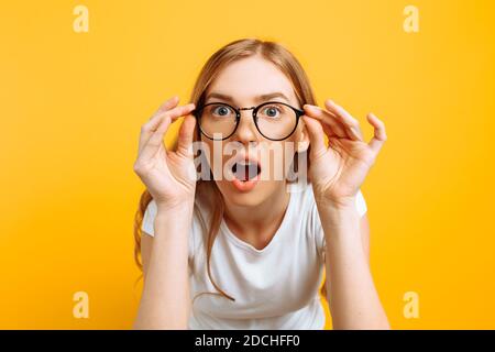 Sorpresa giovane donna che indossa occhiali, indossando una T-shirt bianca, è sorpreso di guardare la fotocamera su uno sfondo giallo Foto Stock