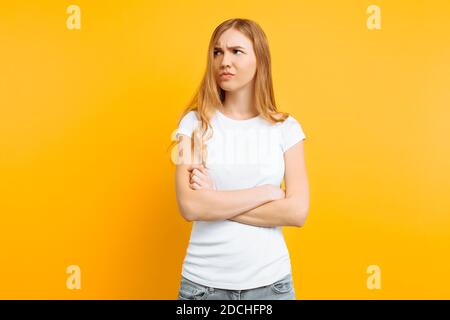 Ritratto di una giovane ragazza capovolto, in una T-shirt bianca, in piedi con le mani piegate e il volto offeso, su uno sfondo giallo Foto Stock