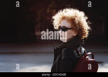 Hipster giovane donna con occhiali da sole e capelli ricci sole. Modello di moda ragazza urbana trendy all'aperto. Stile casual. Gioventù, stile di strada, stile di vita, concetto di tempo libero. Tonificante nello stile degli anni novanta Foto Stock