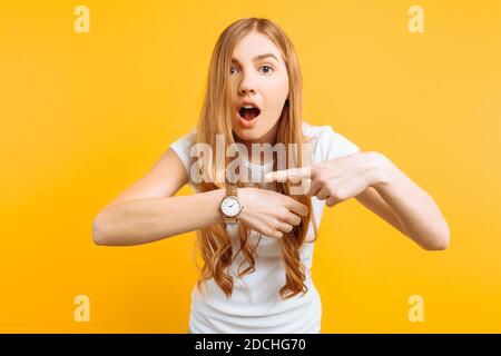 bella ragazza che guarda un orologio a mano, punta all'orologio, su uno sfondo giallo, il concetto di essere in ritardo Foto Stock