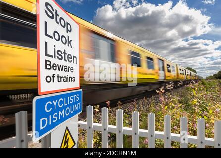 Treno Merseyrail che passa un pubblico a piedi attraversando a velocità con sfocatura del movimento con il segnale di avviso stop look listen e. i ciclisti smontano il segno Foto Stock
