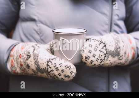 Mani in mittenti caldi che tengono una tazza di caffè grigia con cuore Foto Stock