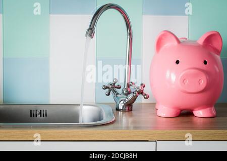 Acqua di rubinetto fluente e una banca di piggy in piedi vicino ad esso. Concetto di prezzo del consumo di acqua Foto Stock