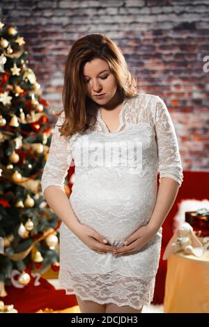 Giovane donna incinta in abito bianco sullo sfondo di decorazioni natalizie Foto Stock