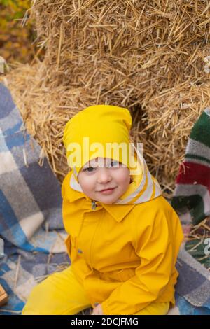 bel ragazzo prescolatore carino in un pantaloni arancione, impermeabile, cappello, stivali in gomma vicino a un mucchio di fieno. Coziness, autunno. Foto Stock