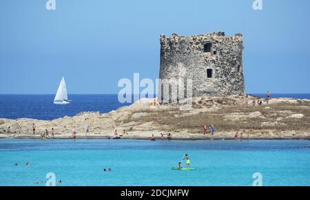 Vista panoramica della bellissima torre la Pelosa con persone in piedi in acqua. Una barca a vela veleggia sullo sfondo Foto Stock