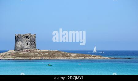 Vista panoramica della bellissima torre la Pelosa con persone in piedi in acqua. Una barca a vela veleggia sullo sfondo Foto Stock