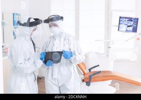 Gli Stomatologi indossano tute ppe che discutono la diagnosi del paziente durante la covid-19. Team di stomatologia in studio dentistico con tuta protettiva agasint coronavirus contagioso durante la pandemia globale. Foto Stock