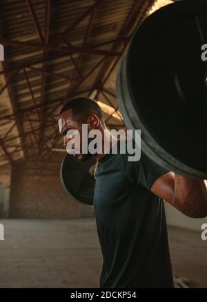 Ma forte esercitarsi a squadre posteriori con un barbell. Uomo che fa squats con pesi pesanti in ombra vecchia fabbrica. Foto Stock