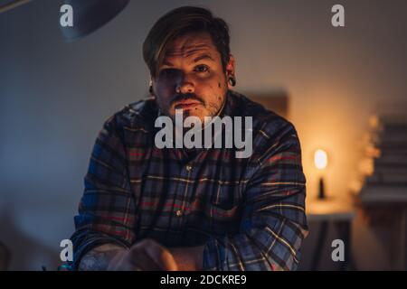 Un ritratto closeup di un uomo hipster serio in un camicia a quadri che guarda la fotocamera mentre sei a casa Foto Stock