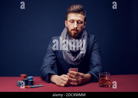 Elegante uomo con bearded in costume e sciarpa che gioca al casinò scuro, fumando sigaro, beve whiskey. Foto Stock