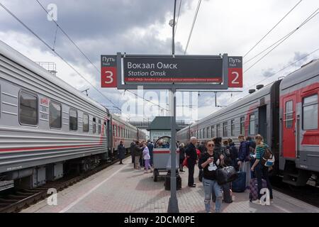 I passeggeri riposano accanto al cartello elettronico sulla piattaforma tra due treni in piedi durante una sosta alla stazione ferroviaria in primavera. Omsk. Foto Stock