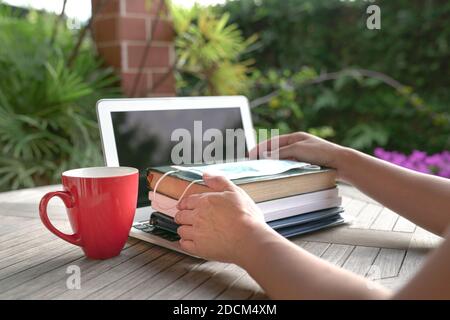 Donna mani su libri impilati con maschera facciale sulla parte superiore del portatile aperto con coppa rossa. Studio a casa durante il concetto pandemico. Foto Stock
