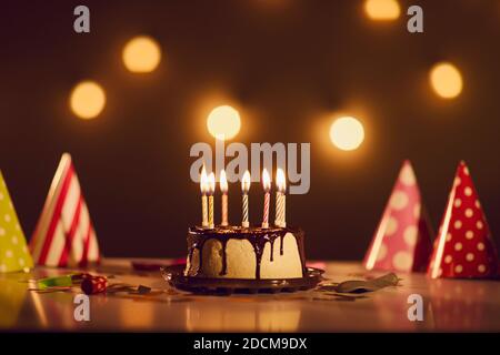 Torta di compleanno con ganache al cioccolato e candele brucianti tavolo con cappelli da festa Foto Stock