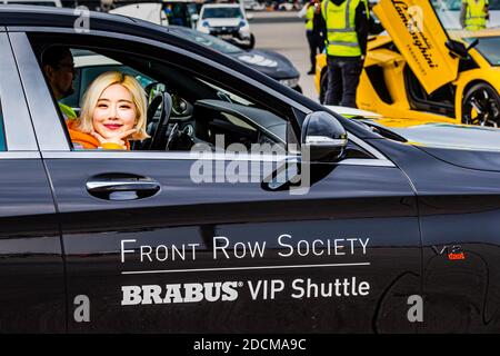 Donna con capelli biondo platino in giubbotto di sicurezza arancione nella navetta VIP Brabus all'aeroporto di Berlino Tegel. Mercedes nera con auto sportiva gialla visibile sullo sfondo della pista dell'aeroporto. Operazioni aeroportuali professionali con veicoli di lusso. Foto Stock